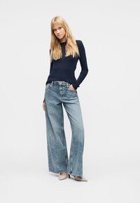 Blond kvinne med langt hår iført en marineblå langermet topp, vide lyseblå jeans og beige spisse hæler, stående med én hånd i lommen.