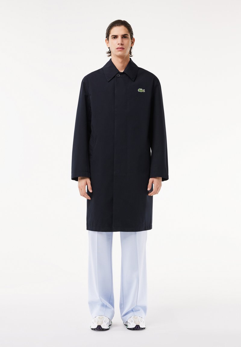 Abrigo azul marino de corte recto, con gran solapa y logo verde; combinado con pantalones de pierna ancha de color azul claro y zapatillas deportivas. Diseño sencillo.