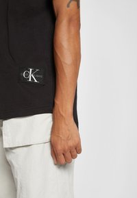 Černé bavlněné tričko s nášivkou loga Calvin Klein Jeans na lemu, částečně viditelné světle zbarvené texturované cargo kalhoty.