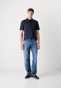 Polo de marinha de mangas curtas, colarinho plano e logótipo no peito; combinado com jeans de perna reta azul claro e sapatos escuros.