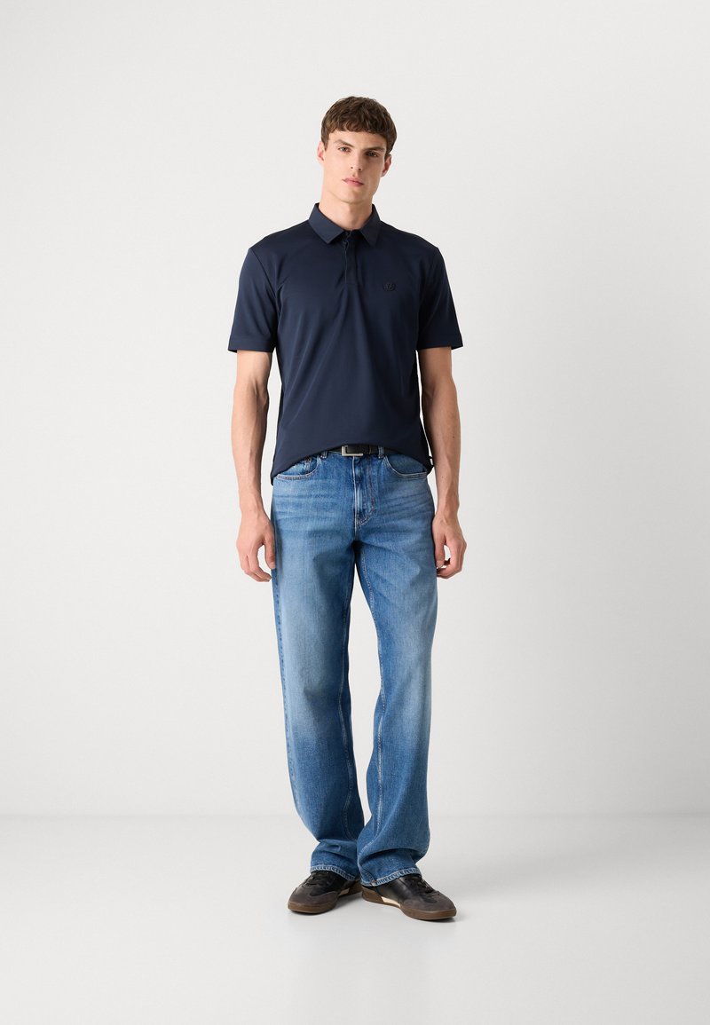 Polo-shirt bleu marine à manches courtes, col plat et logo sur la poitrine ; associé à un jean droit bleu clair et des chaussures foncées.