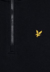 Μαύρος πλεχτός πουλόβερ με μισό φερμουάρ, ριμπ υφή και μικρό κεντημένο κίτρινο έμβλημα αετού στην αριστερή πλευρά του στήθους.