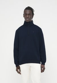 Μπλε-ναυτικό φούτερ με φερμουάρ, σε απαλό ύφασμα, με ψηλό γιακά και ριμπ μανσέτες. Μικρό λογότυπο στο στήθος, φορεμένο με λευκό παντελόνι.