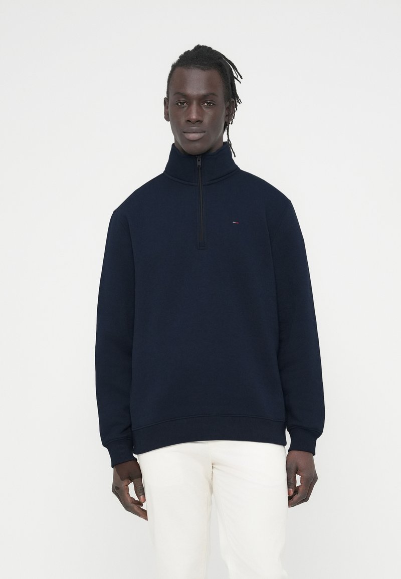 Μπλε-ναυτικό φούτερ με φερμουάρ, σε απαλό ύφασμα, με ψηλό γιακά και ριμπ μανσέτες. Μικρό λογότυπο στο στήθος, φορεμένο με λευκό παντελόνι.