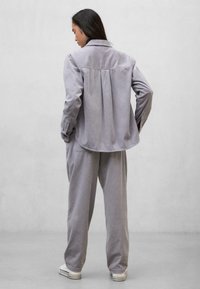 Kvinde der vender væk, iført løs grå corduroy skjorte og bukser med hvide sneakers, stående mod en ensfarvet lys grå baggrund.