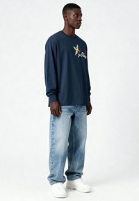 T-shirt à manches longues bleu marine avec une texture douce, arborant un graphisme de chérubin imprimé et le texte  « Je T'aime », associé à un jean en denim bleu clair et des baskets blanches.