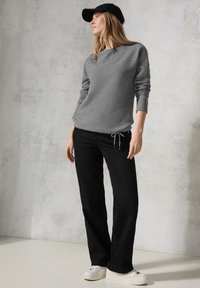 Grijze gebreide sweatshirt met lange mouwen, zwarte drawstring broek en witte sneakers. Model draagt een zwarte pet en poseert tegen een lichte achtergrond.