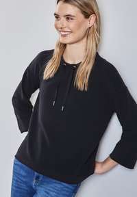 Svart hoodies med ribbad textur, dragsko-detaljer och avslappnad passform. Kombinerad med blå jeans mot en neutral bakgrund.