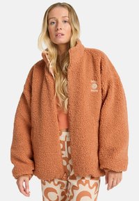 Fleece jack in verbrande sinaasappel, met een hoge kraag en een losse pasvorm. De textuur is pluizig met een logo op de linkerborst. Ritsdetail aan de voorkant.