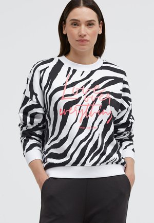 Frau trägt ein schwarz-weiß gestreiftes Zebra-Sweatshirt mit pinkem Text "Love over everything", die Hände in den Taschen einer dunklen Hose.