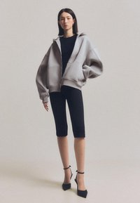 Világosszürke, oversized zipzáros kapucnis felső, lecsúszott vállakkal, egy testhezálló fekete toppal és fekete, térd alá érő leggings-szel, valamint fekete bokapántos magassarkúkkal párosítva.