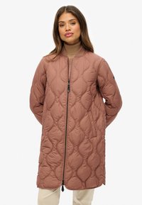 Femme portant une longue veste matelassée rose poussiéreux avec une fermeture éclair et un col côtelé sur un pull à col roulé marron et un pantalon clair.