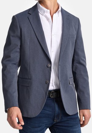 Homme portant un blazer gris sur une chemise blanche boutonnée, un jean bleu foncé et une ceinture noire, debout devant un fond blanc.