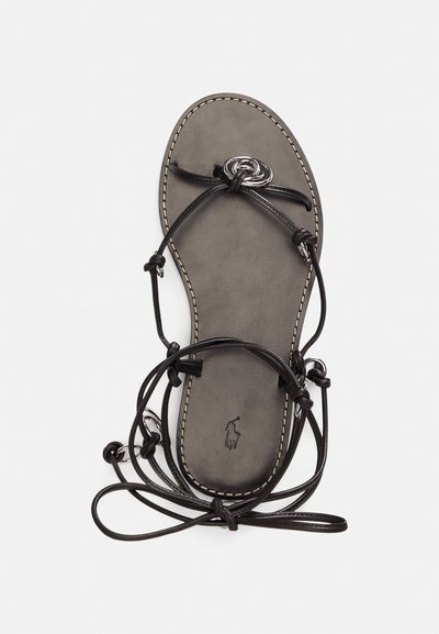 Μαύρο δερμάτινο σανδάλι με λεπτά λουράκια στον αστράγαλο, κυκλικό μεταλλικό διακοσμητικό και γκρι υφασμάτινη εσωτερική σόλα. Διαθέτει ραφές κατά μήκος της άκρης της σόλας.