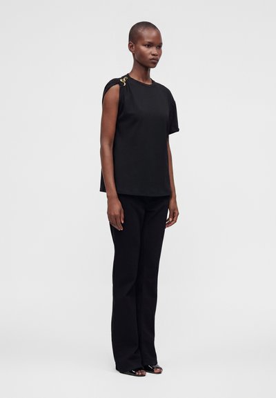 Femme portant un haut noir sans manches avec un fermoir doré sur une épaule, un pantalon noir à jambes larges et des talons ouverts noirs, debout devant un fond uni.