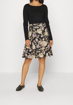 Frau trägt ein schwarzes Langarmoberteil kombiniert mit einem knielangen schwarzen Rock mit großem rosa-beigem Blumenmuster.