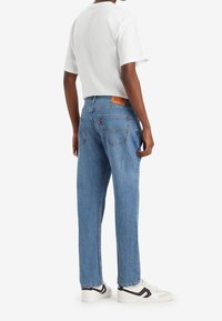 Ljusblå denimjeans med avslappnad passform, femficksdesign och en läderlapp på midjebandet, bärs tillsammans med en vit t-shirt och sneakers.