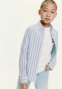 Cămașă ușor albastră cu dungi verticale roșii, purtată peste un tricou alb, asociată cu blugi de denim deschis. Aspect casual și confortabil.