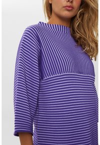 Frau mit einem lila und lavendelfarben gestreiften Pullover, der unterschiedliche horizontale und vertikale Streifenmuster aufweist, und schulterlangen blonden Haaren.