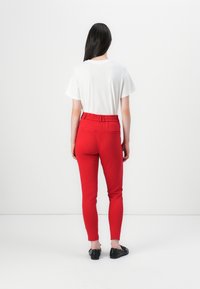 Une personne avec de longs cheveux noirs se tient de dos, portant un t-shirt blanc et un pantalon rouge, contre un fond blanc uni.