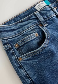 Jeans drepti de gleznă din denim albastru, cu talie medie, buzunar frontal vizibil cu nituri și buclă pentru curea, etichetă care indică fabricat în Pakistan.