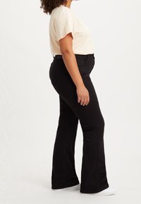 Jeans noirs évasés en denim, avec une texture lisse. Assortis à une chemise à manches courtes beige clair. Les chaussures sont blanches avec un design simple.