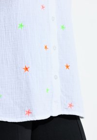 Weißes, strukturiertes Stoffhemd mit neon-grüner, oranger und pinker Sternstickerei sowie weißen Knöpfen über schwarzer Hose.