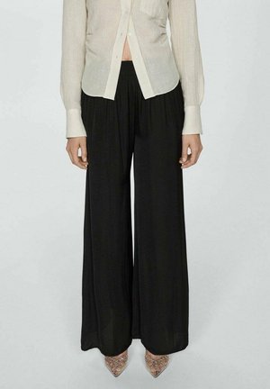 Osoba ubrana w przezroczystą białą bluzkę na guziki, czarne szerokie spodnie z wysokim stanem oraz przezroczyste szpilki z ozdobami.