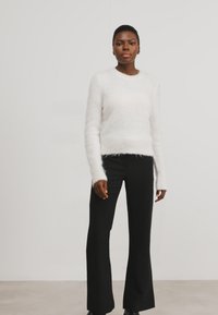 Pullover bianco morbido con scollatura rotonda e maniche lunghe, abbinato a pantaloni neri a palazzo. Sfondo neutro.