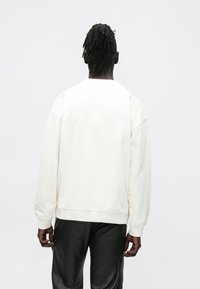 Λευκό φούτερ με στρογγυλή λαιμόκοψη και χαλαρούς ώμους, με ριμπ μανσέτες και τελείωμα, φτιαγμένο από απαλό βαμβακερό ύφασμα, φορεμένο με μαύρο παντ�ελόνι.