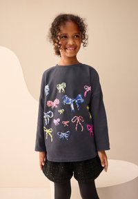 Temno siva dolg rokav s potiski barvnih pentlji iz sequins, v kombinaciji s črnimi teksturiranimi kratkimi hlačami. Gladek tekstil, sproščen kroj.