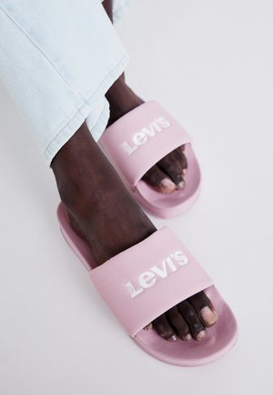 Chanclas de color rosa claro con una correa ancha que presenta la marca "Levi's" en blanco; material de goma suave; diseño de punta abierta, llevadas con jeans de color azul claro.