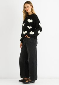 Black sweater with white heart patterns, long sleeves, and textured knit. Paired with black wide-leg jeans and black flats.