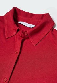 Polo shirt rouge avec un col, présentant une patte de boutonnage avec deux boutons assortis. Fabriqué en tissu léger et lisse. Étiquette visible à l'intérieur du col.