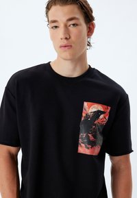 Camiseta de algodón negra de mangas cortas y cuello redondo, con un estampado gráfico en rojo y negro de un personaje frente a una luna llena.