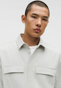 Hele hall särgi, millel on kaks rinna taskut, struktuurne krae ja sile tekstuur. Kantud valge T-särgi peal.