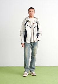Lett, glidelåsjakke i lys grå med marineblå detaljer, parret med lyseblå jeans og brune og hvite joggesko. Jevn tekstur, avslappet passform.