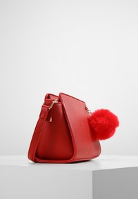 Czerwona torebka z syntetycznej skóry o zwartej formie, z dużym pomponem z czerwonego sztucznego futra oraz złotymi elementami ozdobnymi.