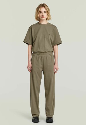 Femme aux cheveux blonds courts, debout, portant une chemise ample à manches courtes vert olive avec un pantalon assorti et des chaussures noires sur un fond clair.