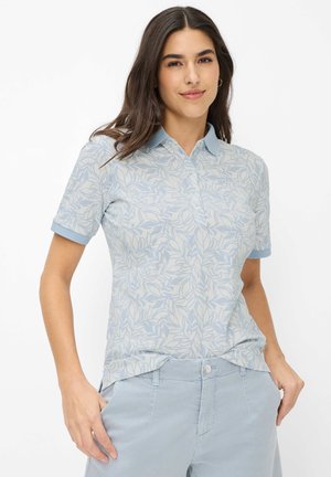 Kvinde med langt mørkt hår iført en lyseblå polo med bladmønster og matchende lyseblå bukser, hænderne i lommerne, smilende let.