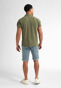 Camisa polo verde aceituna de manga corta, combinada con pantalones cortos azul claro enrollados y zapatillas crema. Diseño sencillo con un ajuste casual.