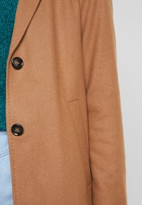 Brązowy płaszcz wełniany z domieszką, z dwoma czarnymi guzikami i boczną kieszenią, nałożony na niebieską sweterek o teksturowanej fakturze.