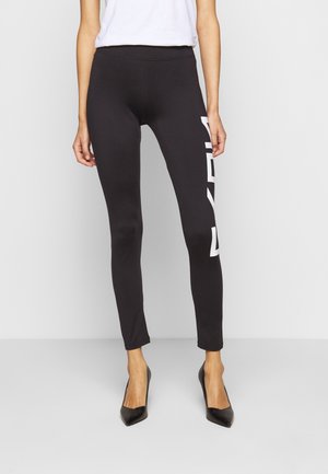 Vrouw in zwarte slim-fit leggings met witte zijprint en zwarte puntige hakken, staand tegen een effen lichte achtergrond.