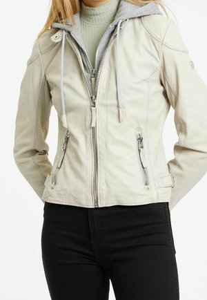 Frau trägt eine hellbeige Jacke mit Reißverschluss, Reißverschlusstaschen und einer grauen Kapuze über einem grünen gerippten Rollkragenpullover und schwarzen Hosen.