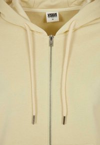 Sweat à capuche crème avec fermeture éclair, cordons, fermeture éclair métallique et étiquette « Urban Classics Organic » en taille XS à l'intérieur du col.