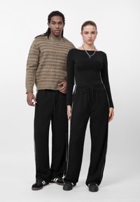 Két modell laza öltözéket visel: egy csíkos kötött pulóvert és egy testhezálló fekete felsőt, fekete melegítőnadrággal, fehér oldalsávokkal.