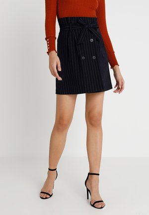 Femme portant une jupe portefeuille noire à fines rayures avec des boutons et une ceinture à nouer, associée à un top orange à manches longues côtelé et des sandales à talons noirs.