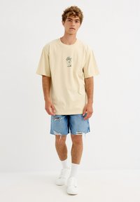 Joven de pie con camiseta beige oversize con gráfico, pantalones cortos de mezclilla rotos, calcetas blancas y zapatillas blancas, sobre fondo blanco liso.