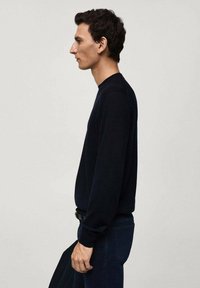 Maglione navy a maniche lunghe e collo rotondo, abbinato a jeans scuri. Il tessuto appare liscio e leggermente aderente, con dettagli minimi.
