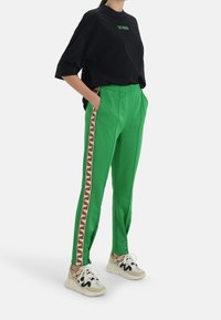 Gröna träningsbyxor med en brun zigzag-rand längs sidan, kombinerat med en svart t-shirt och beiga sneakers med svarta detaljer.